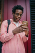 © BullRun - Young African man in glasses smiling at smartphone while leaning on red wall, capturing digital relaxation, joyful tech usage and ease of mobile connection in daily freelance flow.