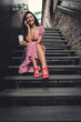 © BullRun - Smiling Hispanic woman in pink dress with curly hair sits on stairs, coffee and smartphone in hand, enjoying remote work freedom and digital communication in casual lifestyle.