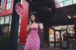 © BullRun - Confident Hispanic woman in pink floral dress with smartphone and coffee cup smiles while walking outdoors, reflecting mobile communication, freelance lifestyle, and joyful independence.