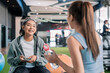 © whyframeshot - Two cheerful Asian women chatting and laughing together at a gym, one holding a smartphone while talking to her friend, enjoying a fun break between workouts.