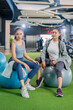 © whyframeshot - Two attractive Asian women resting together at a modern gym, sitting on exercise balls with water bottles, wearing colorful sportswear in a bright fitness club.