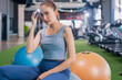 © whyframeshot - confident Asian woman sitting on exercise balls at a modern gym, wearing a gray sports top and blue leggings with white sneakers, posing with a calm and focused look.