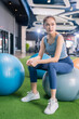 © whyframeshot - confident Asian woman sitting on exercise balls at a modern gym, wearing a gray sports top and blue leggings with white sneakers, posing with a calm and focused look.