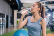 © whyframeshot - smiling Asian woman resting at the gym, holding a water bottle while sitting on an exercise ball, wearingactive sport cloth leggings in a modern fitness club.