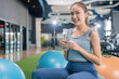 © whyframeshot - smiling Asian woman resting at the gym, holding a water bottle while sitting on an exercise ball, wearingactive sport cloth leggings in a modern fitness club.