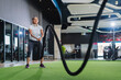 © whyframeshot - fit Asian woman doing battle rope exercise at a modern gym, holding thick black ropes in a wide stance, wearing sport active cloth leggings with intense focus.