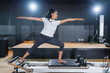 © whyframeshot - Asian woman doing Pilates reformer exercise in a studio, kneeling and pulling resistance band with focused concentration, wearing white top and black leggings.