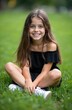© miss irine - Young girl with long brown hair smiles sitting cross-legged on green grass. Wears black off-shoulder dress, white sneakers, enjoying summer day outdoors with carefree happy expression. Image ideal