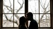 © Sophia - Romantic couple kissing in dark silhouette stands together in a tender embrace in front of a large bright window with a view of bare trees outside, creating a moody scene