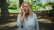 © Krakenimages.com - Senior woman holding cheek with hand in forest park, eyes closed and wincing from toothache while wearing light blue blouse and standing on shaded path; pain vulnerability.