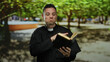 © Krakenimages.com - Hispanic priest reading bible outdoors in a sunny park, wearing black robe and cross, surrounded by trees.
