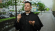 © Krakenimages.com - Priest holding cross on urban street looks contemplative under trees with blurred cityscape in background.