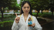 © Krakenimages.com - Young asian woman doctor in uniform with stethoscope points at medicine bottle in a lush green park setting.