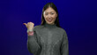 © Krakenimages.com - Asian woman pointing aside against a solid blue background wearing a grey sweater, conveying direction or suggestion with a confident expression.