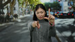 © Krakenimages.com - Woman gesturing loser sign outdoors on urban street with confident expression pointing forward, wearing gray sweater, standing amid city surroundings on a bright day.