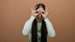 © Krakenimages.com - Woman posing playfully with hands making binoculars over eyes against isolated brown wall background, showcasing confident joyful expression with stylish long hair.
