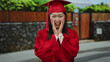 © Krakenimages.com - Woman in red graduation gown celebrating outdoors on urban street with joyful expression, surrounded by greenery and city elements, showcasing pride and success.