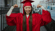 © Krakenimages.com - Woman in red graduation gown flexing muscles proudly in an urban street setting, symbolizing achievement and success.