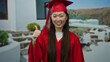 © Krakenimages.com - Woman in red graduation gown outdoors smiling and pointing up at scenic backdrop with white walls and natural landscape visible in the background.