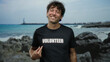 © Krakenimages.com - Hispanic man smiling on the beach points to his volunteer shirt with the sea and rocks in the background, conveying community spirit and environment awareness.