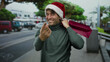 © Krakenimages.com - Hispanic man in christmas hat smiling and gesturing come with finger while holding shopping bag on a street with blurred cars and trees.