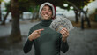 © Krakenimages.com - Hispanic man wearing christmas hat smiles holding us dollars on an outdoor street, displaying joy and holiday spirit.