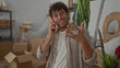 © Krakenimages.com - Hispanic man talking on phone in new home surrounded by moving boxes and plants, smiling confidently while gesturing to emphasize his conversation.