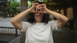 © Krakenimages.com - Hispanic middle-aged woman playfully mimicking binoculars with her hands outdoors, smiling in a casual setting with greenery and architectural elements in the background.