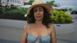 © Krakenimages.com - Middle-aged hispanic woman in bikini and sun hat stands outdoors on a sunny street with flowers and buildings in the background, showcasing a relaxed summer vibe.
