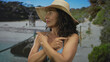 © Krakenimages.com - Middle-aged hispanic woman in blue bikini and hat on sunny beach, exuding confidence and relaxation by the seaside with natural greenery in the background.