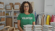 © Krakenimages.com - Middle-aged hispanic woman volunteer in green uniform working in charity indoor room surrounded by donation boxes and meal trays, showing appreciation.