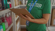© Krakenimages.com - Woman wearing volunteer uniform checks inventory on clipboard in indoor charity room filled with hygiene products on shelves, showcasing organization and community service.