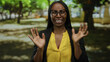 © Krakenimages.com - Woman with glasses and curly hair makes surprised expression in a park setting, wearing a yellow shirt and black jacket, surrounded by blurred green trees and sunlight.