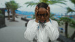 © Krakenimages.com - Doctor stands stressed with curly hair by the seaside in a white coat and glasses, holding her head outdoors with palms in background.