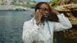 © Krakenimages.com - Woman talking on smartphone at seaside with frustration, wearing glasses and doctor's coat, capturing emotions in a scenic beach setting during daytime.