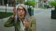 © Krakenimages.com - Senior woman with grey hair and glasses giving thumbs up and listening gestures on a city street, expressing curiosity and positivity outdoors with blurred background.