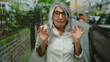 © Krakenimages.com - Senior grey-haired woman with glasses making playful gesture on a city street, blending seriousness with humor, against a backdrop of urban greenery and parked cars.
