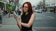 © Krakenimages.com - Woman using smartphone on bustling city street with red hair and glasses overlooking busy urban setting with buses and people.