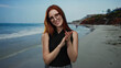 © Krakenimages.com - Smiling redhead woman with glasses applauding at the seaside, capturing the joy of a sunny beach day with waves and distant cliffs in the serene background.