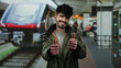 © Krakenimages.com - Hispanic man smiling at a train station, giving a thumbs-up gesture, conveying positivity and travel readiness with a modern railway and passengers in the background.