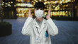 © Krakenimages.com - Young man wearing mask and lab coat adjusting his glasses outdoors in an urban evening setting with blurred lights in the background.
