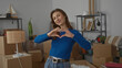 © Krakenimages.com - Woman making heart gesture with hands in new home living room surrounded by boxes, wearing blue sweater, symbolizing happiness and warmth in a fresh start indoors.