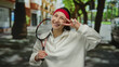 © Krakenimages.com - Woman smiling in city street holding tennis racket making peace gesture wearing white sweater and red headband outdoors surrounded by urban landscape.