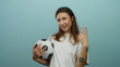 © Krakenimages.com - Woman holding soccer ball showing peace sign against blue wall expressing cheerful emotion and confidence.
