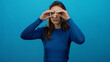 © Krakenimages.com - Woman peering through fingers on isolated blue background suggests curiosity, with a caucasian young lady in a blue sweater embodying inquisitive exploration.
