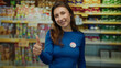 © Krakenimages.com - Young woman in a supermarket wearing a vote badge smiles and gives a thumbs up, emphasizing civic engagement in a commercial indoor setting with colorful shelves behind.