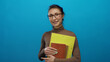 © Krakenimages.com - Woman smiling and holding folders against an isolated blue background in a casual setting, showcasing confidence and style.
