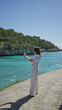 © Krakenimages.com - Woman capturing ocean view on smartphone at sunny beach in santanyi, mallorca with vibrant turquoise water and lush greenery under clear blue sky.