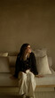 © Krakenimages.com - Woman sitting indoors on sofa in mediterranean home with beige wall and cushions, wearing glasses and sweater, smiling comfortably with natural light filtering in.