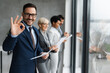 © Novak - Confident businessman standing by office window showing OK gesture.